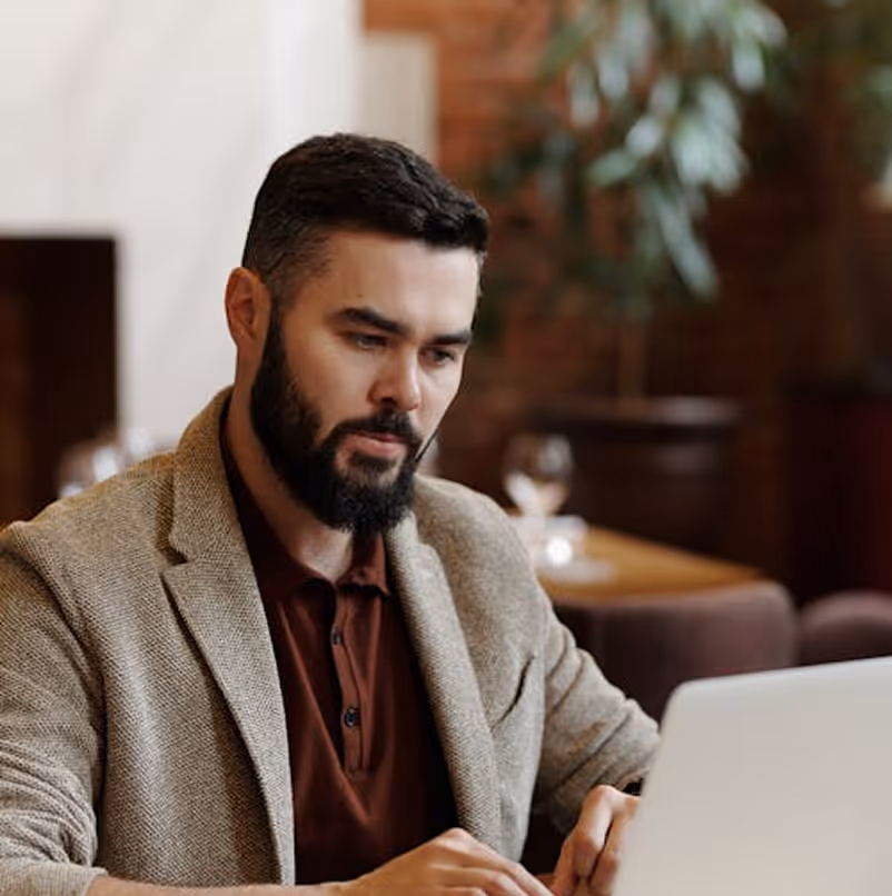 Bearded man in beige blazer working on a laptop in a cozy indoor setting.