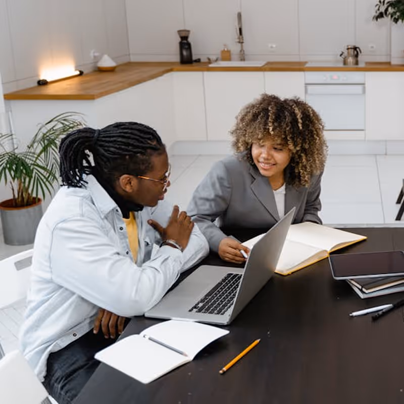 Two people sitting at a table with a laptop, notebook, and tablet, engaged in a discussion in a modern kitchen setting.