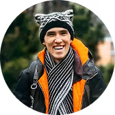 Smiling young man outdoors wearing a black and white patterned beanie, striped scarf, and orange-lined jacket.