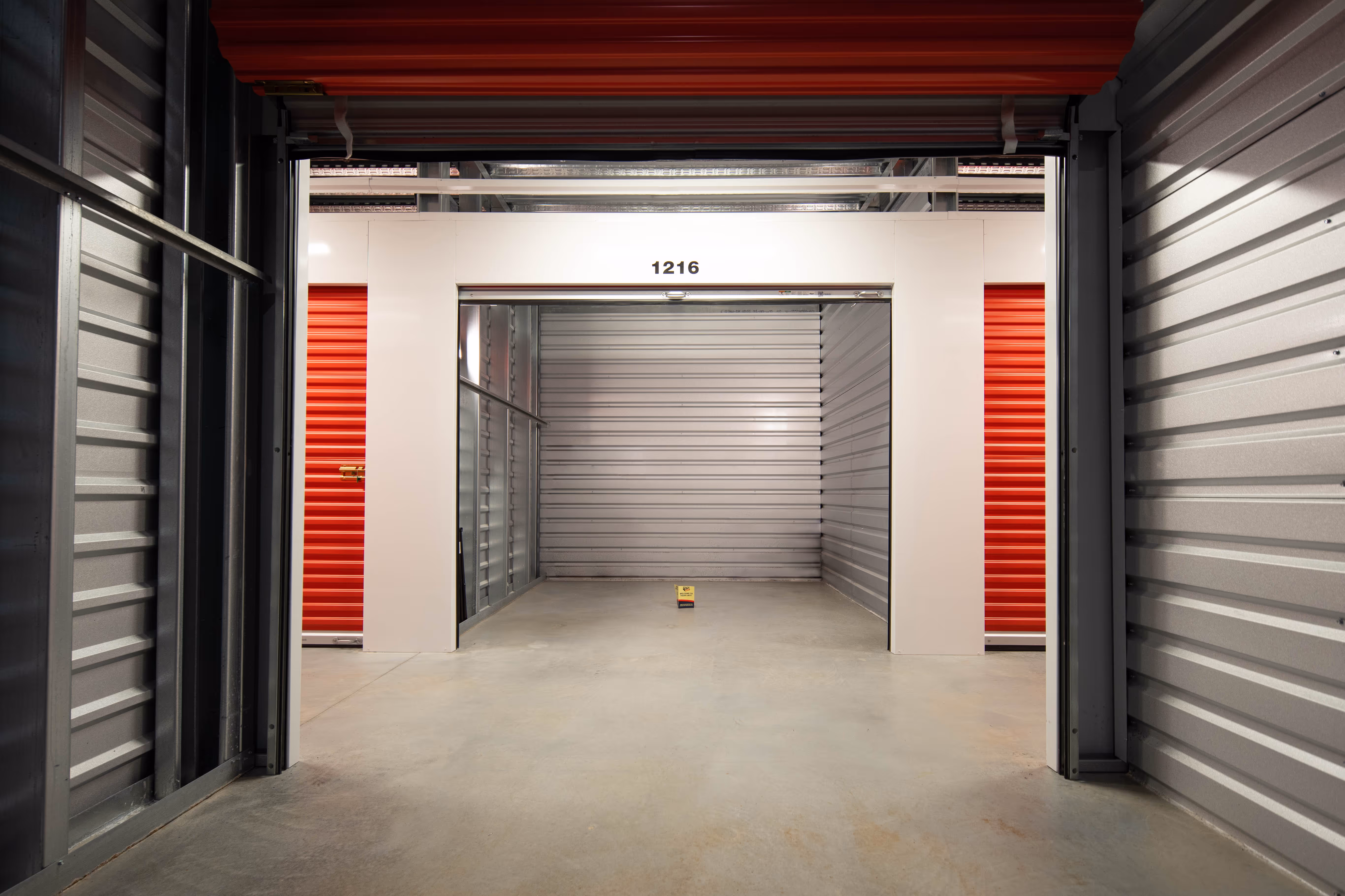 Pack N Stor Self-Storage indoor storage unit with open red roll-up door labeled 1216 and concrete floor.