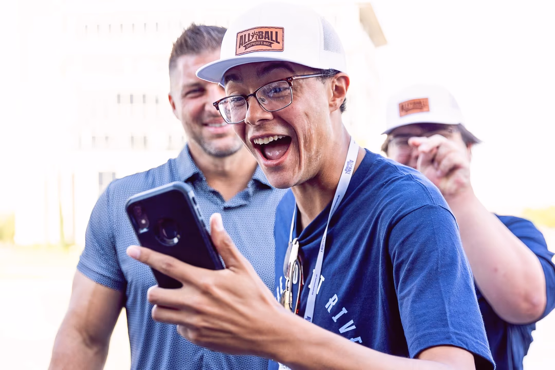 Young man in cap and glasses excitedly looking at a smartphone with two smiling men behind him.