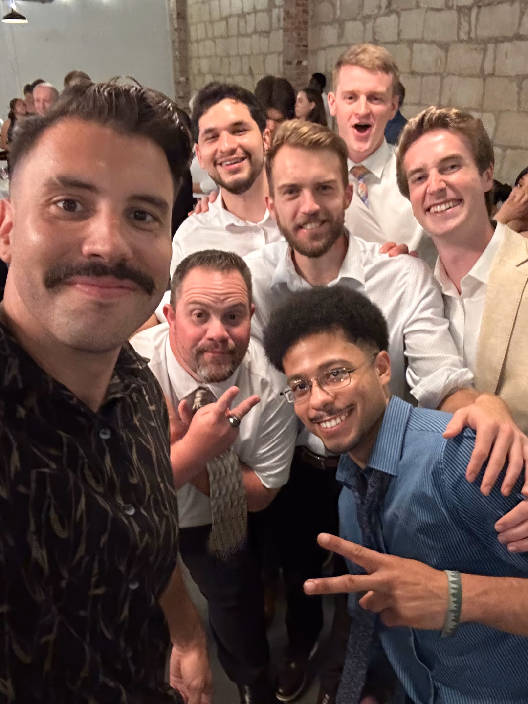 Group of seven men smiling and posing together indoors, some making peace signs.