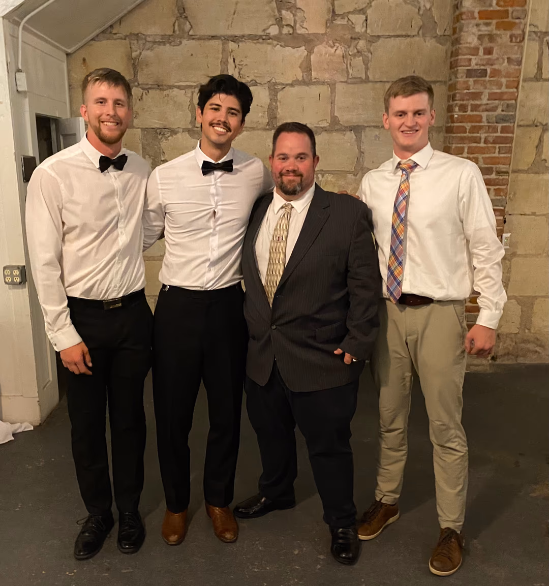 Four men smiling dressed formally, three in white shirts and bow ties or tie, one in dark suit.