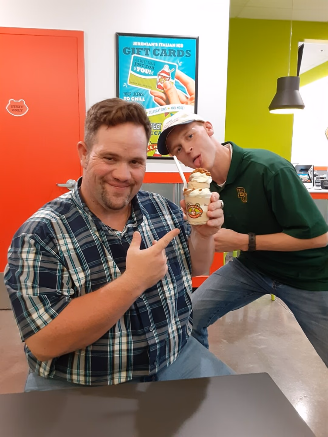 Two men in a colorful room; one points to a dessert cup while the other leans in to lick it.
