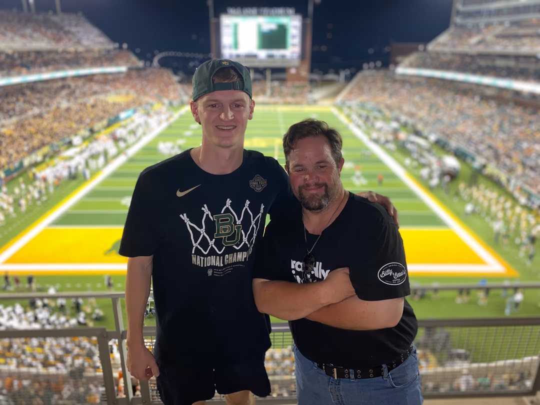 Two men posing and smiling at a brightly lit football stadium during a game.