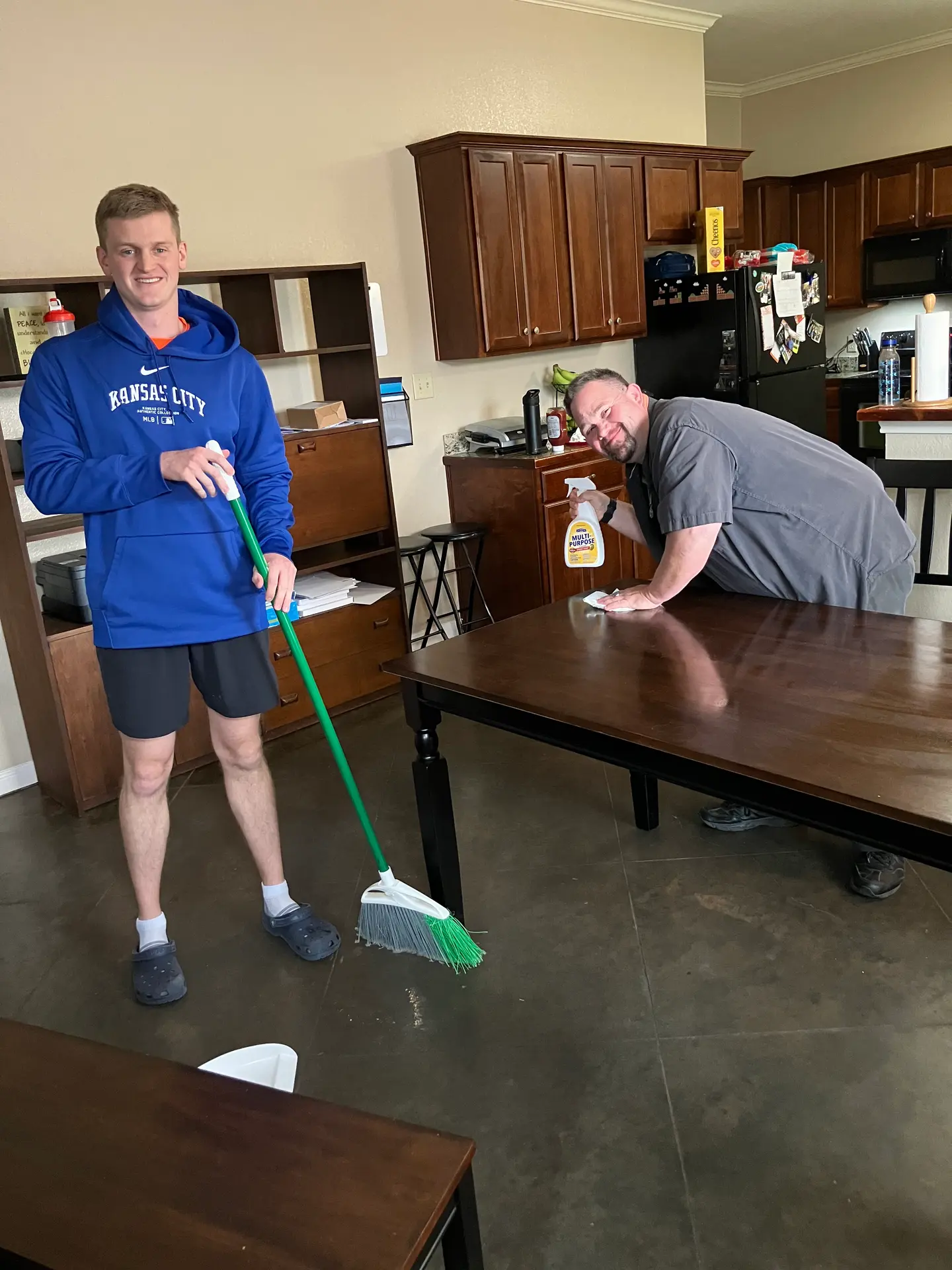Two men cleaning a kitchen: one sweeping floor, the other wiping a wooden table with spray bottle.