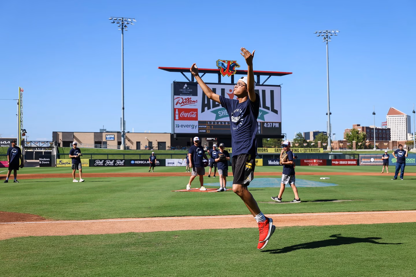 Young man jumping with arms raised on baseball field with other players and scoreboard behind him.