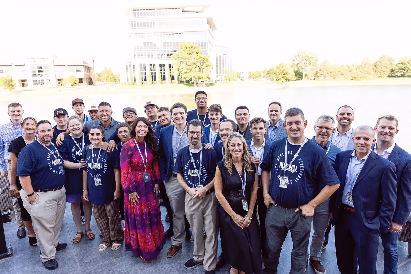 Group of people smiling outdoors by a lake with buildings and trees in the background.