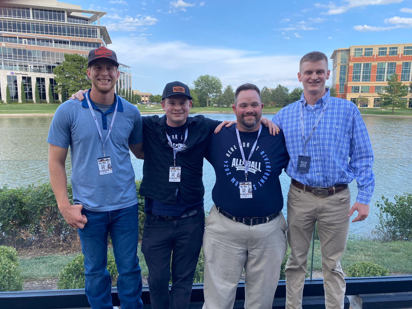 Four men smiling with arms around each other, standing outdoors near a pond with office buildings behind.