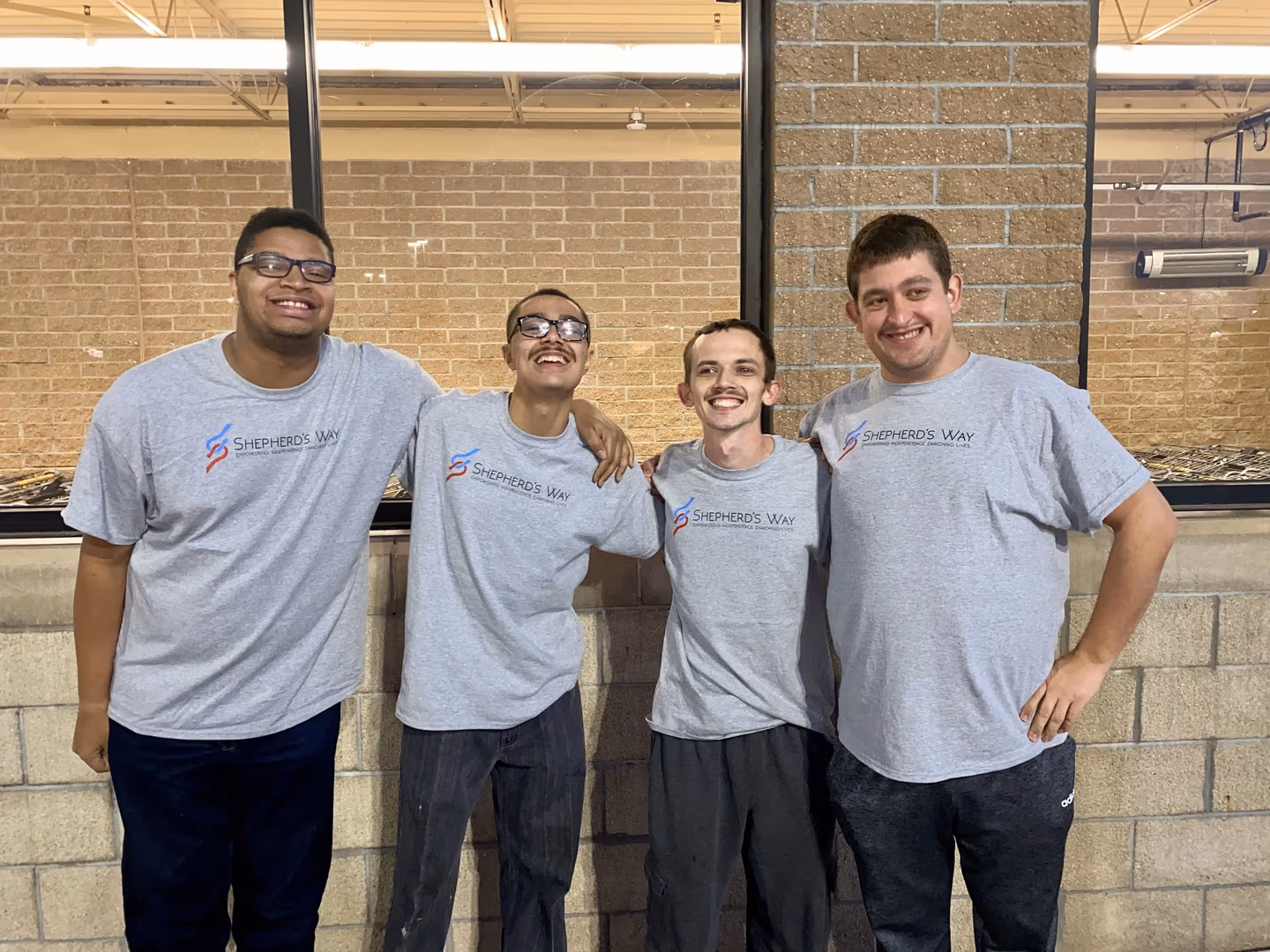 Four young men smiling, wearing matching Shepherd's Way gray T-shirts, standing linked arm in arm.