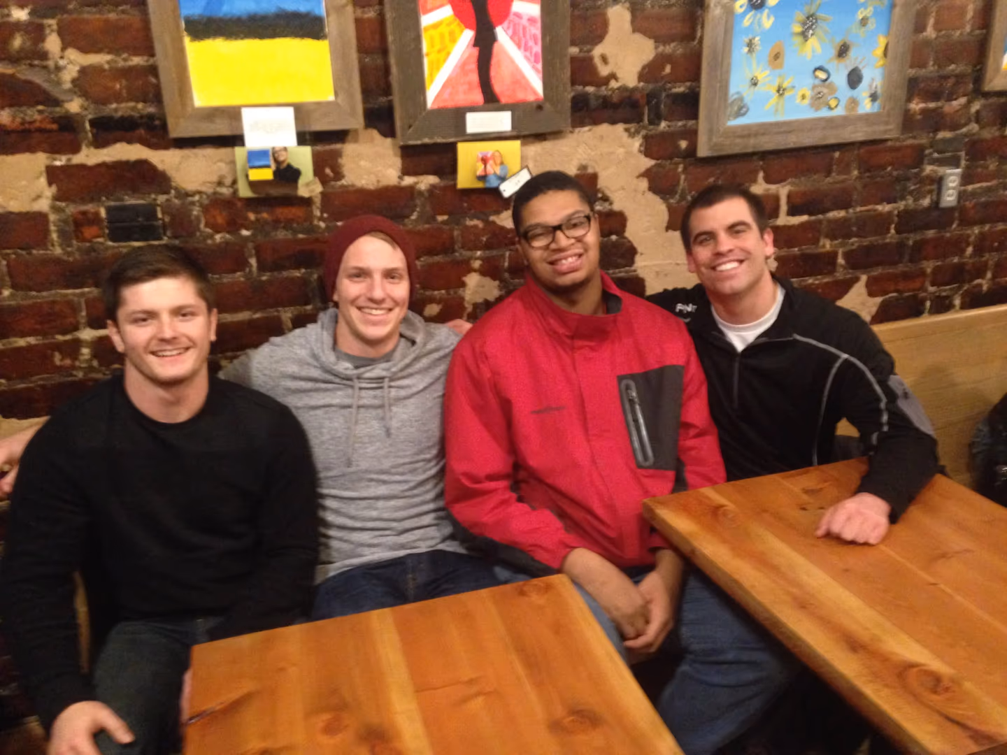 Four young men smiling and sitting at wooden tables with art on a brick wall behind them.