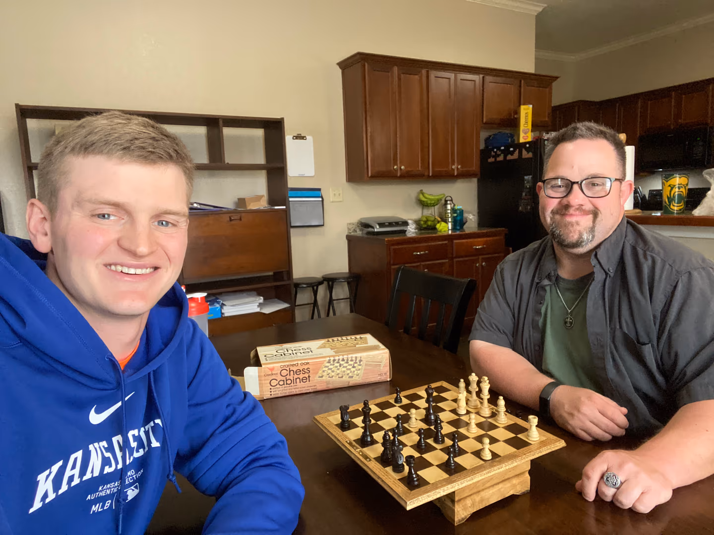 Two men sitting at a table playing chess with a wooden chessboard and pieces.