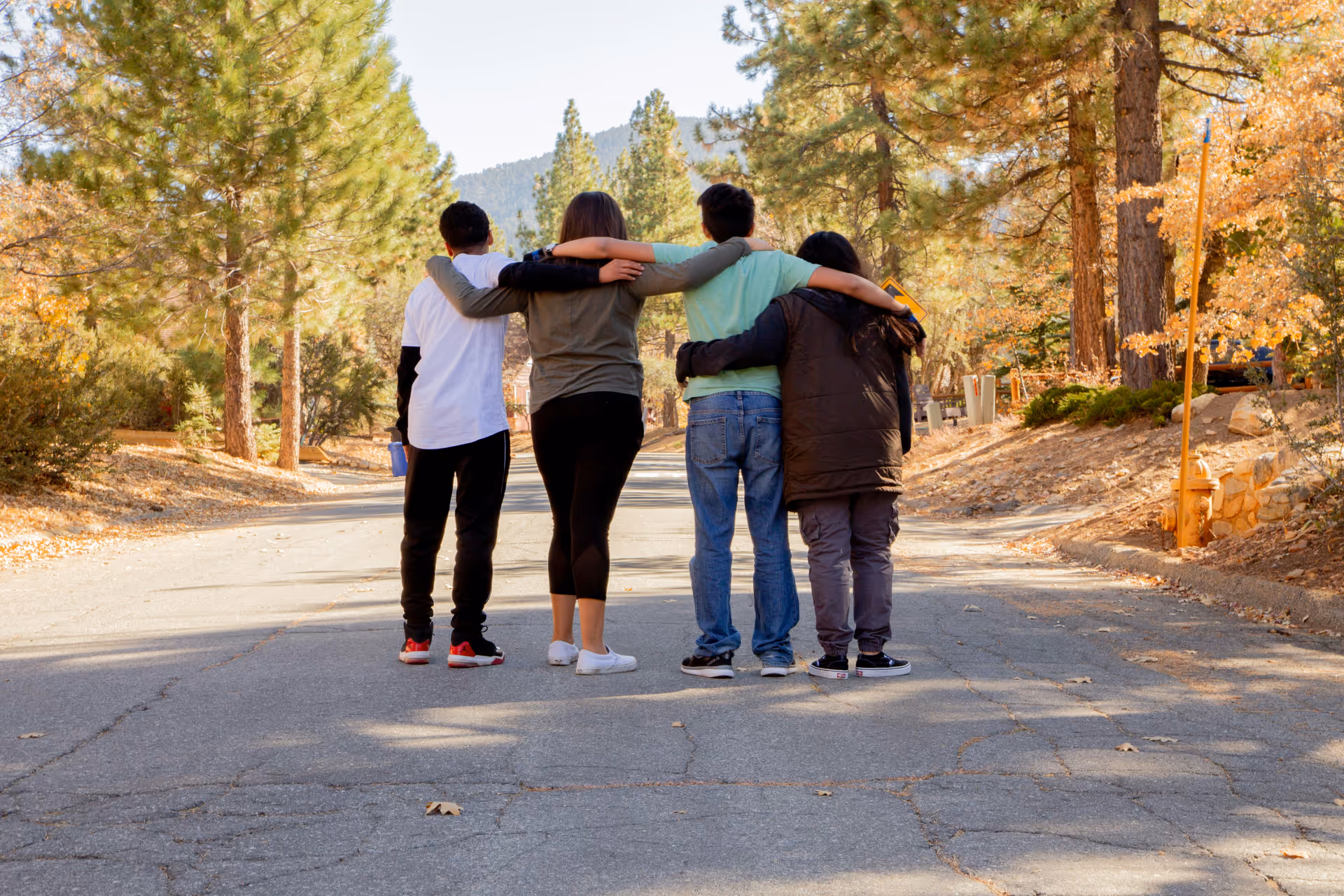 Four young friends with arms around each other standing on a tree-lined street in autumn.