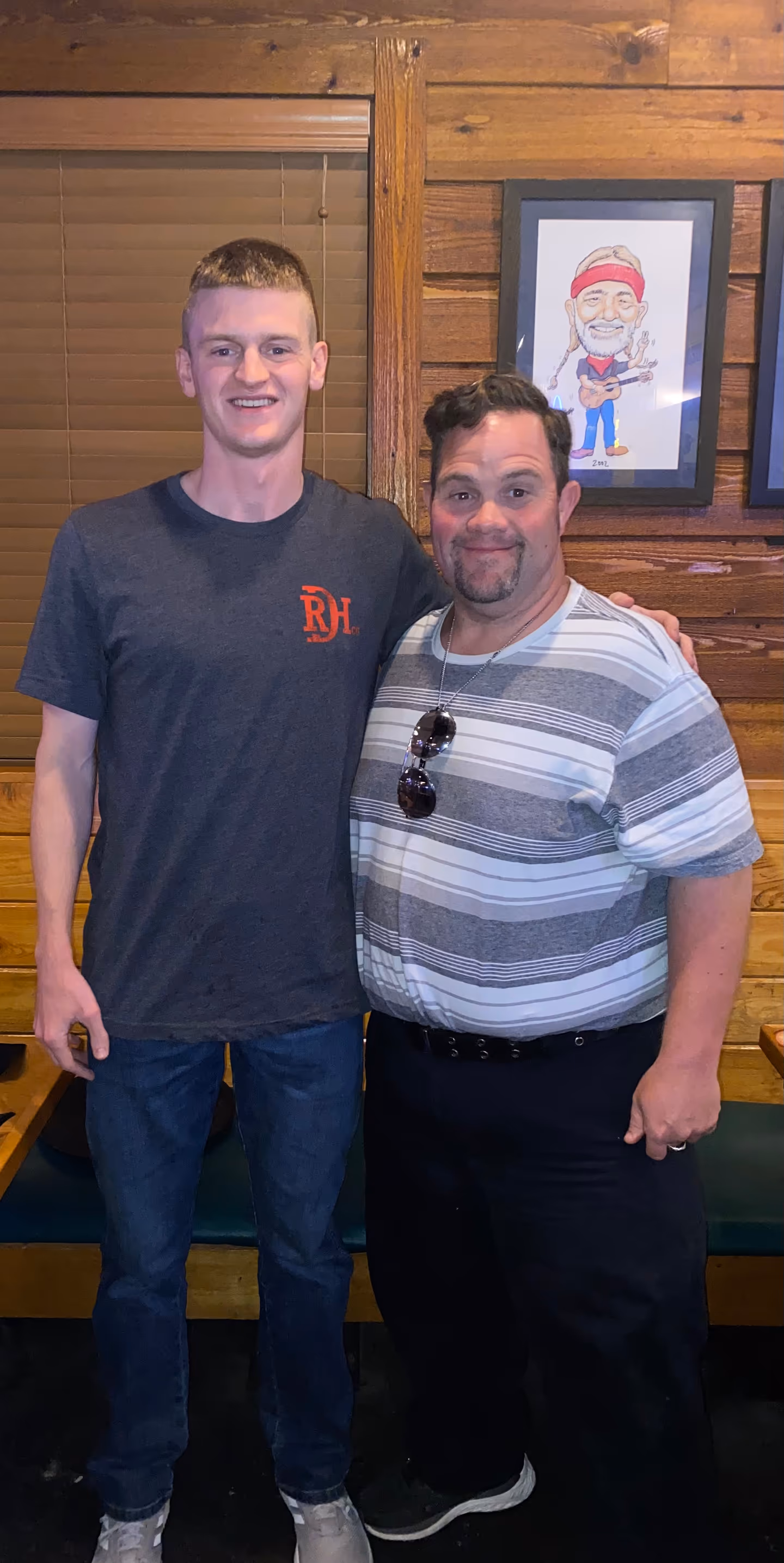 Two men smiling, one wearing a dark t-shirt and jeans, the other in a striped shirt with sunglasses.