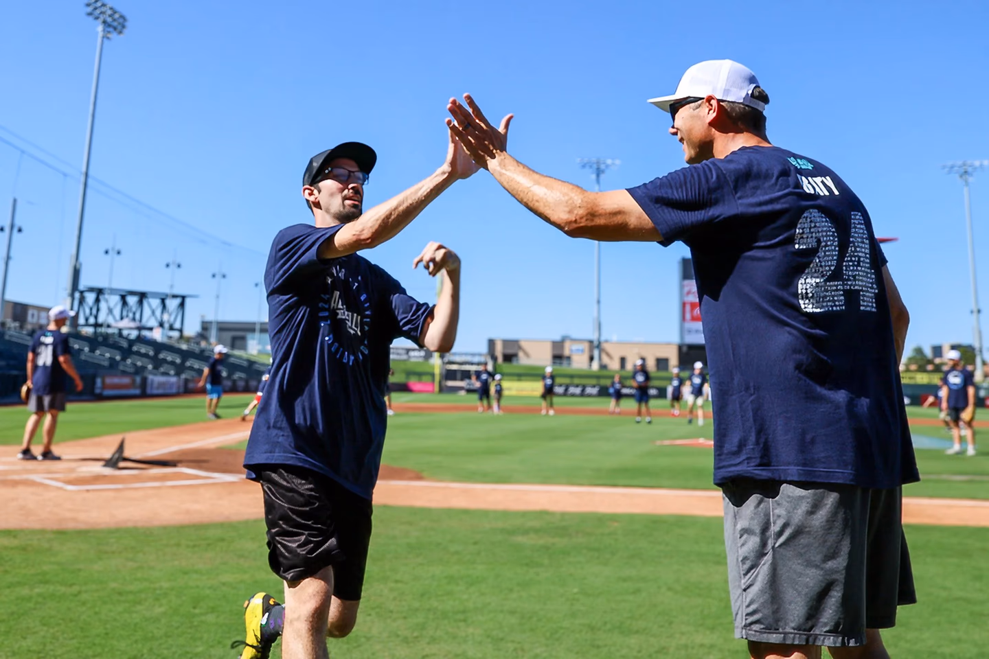 Two men in sportswear giving a high five on a baseball field with players in the background.