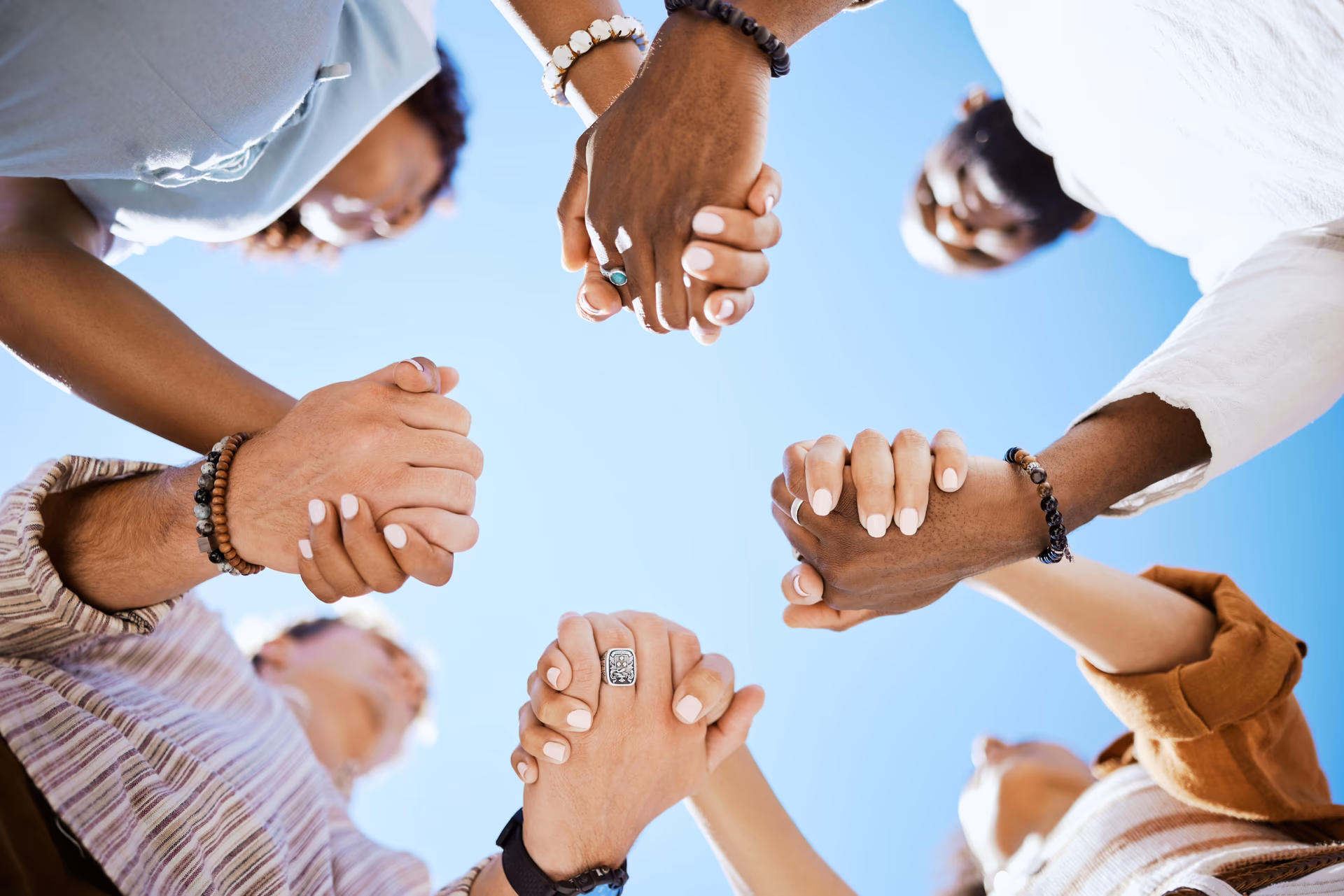 Group of diverse people standing in a circle holding hands against a clear blue sky.