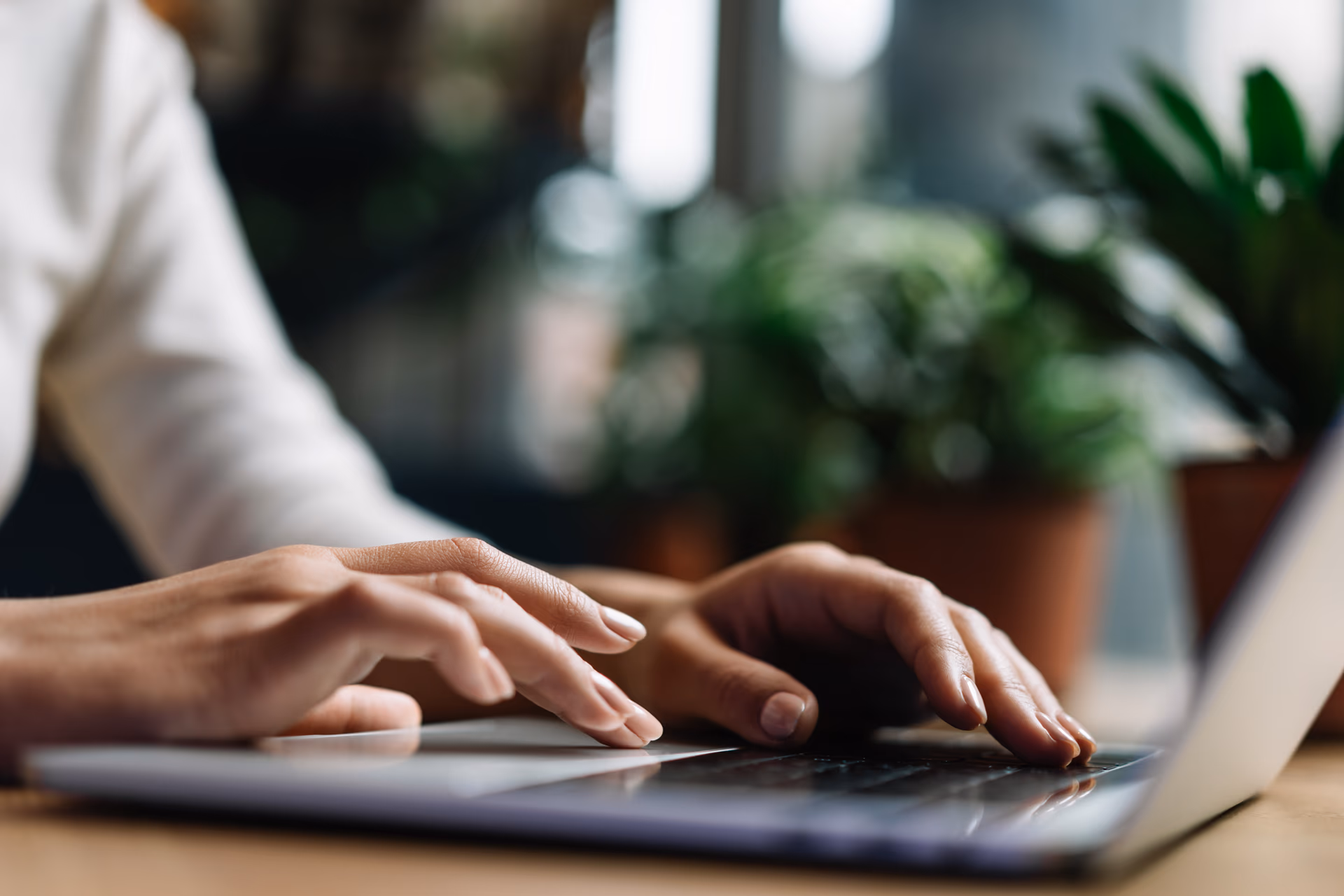 Close-up of hands typing on a laptop keyboard with blurred plants in the background.