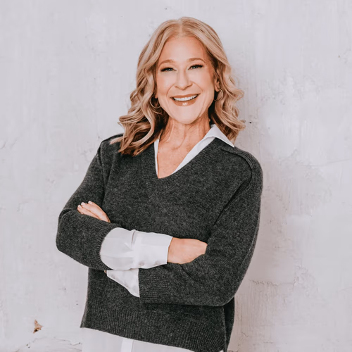 Smiling woman with blonde wavy hair in a dark sweater and white shirt, arms crossed, against a white wall.