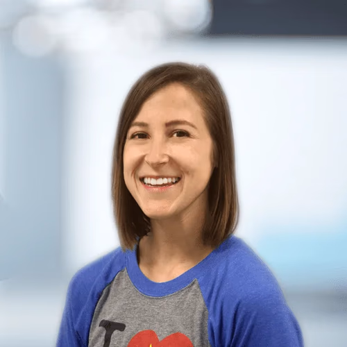Smiling woman with straight brown hair wearing a blue and gray shirt in a blurred indoor setting.