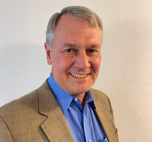 Smiling middle-aged man wearing a blue shirt and brown blazer against a plain light wall.