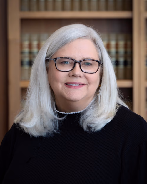 Smiling woman with white hair and glasses standing in front of a bookshelf with law books.