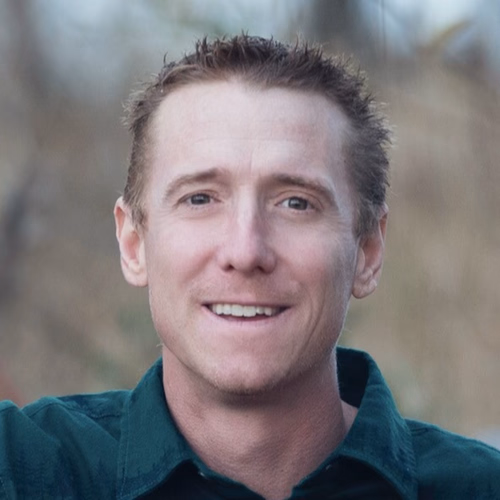 Smiling man with short brown hair wearing a dark green collared shirt outdoors.