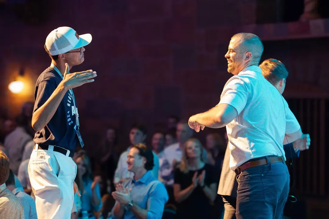 Two men facing each other with one gesturing and the other smiling, audience clapping in background.