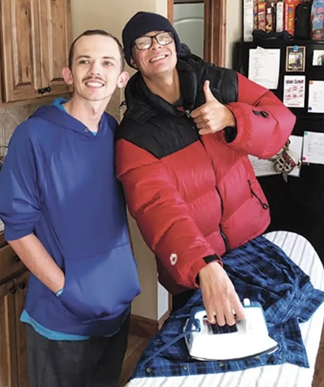 Two smiling men in a kitchen, one ironing a blue plaid shirt while the other stands nearby.