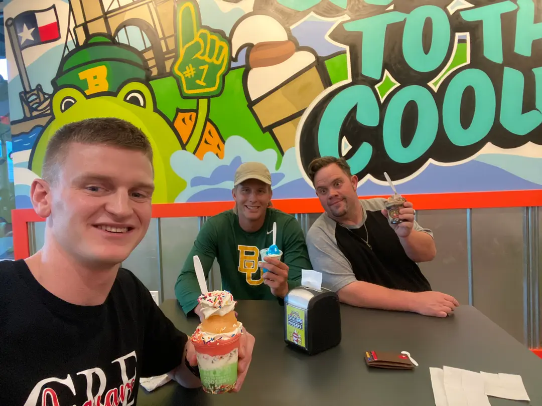 Three men smiling with ice cream cups seated at a table in front of a colorful mural.