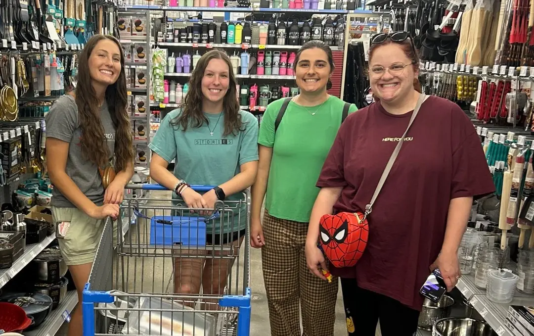 Four smiling women standing in a store aisle with a shopping cart and various products on shelves.