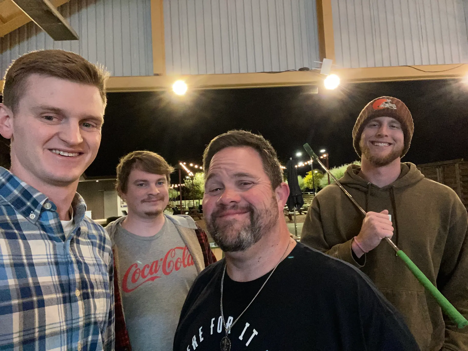 Four men smiling outdoors at night, one holding a green-handled golf club, under string lights.