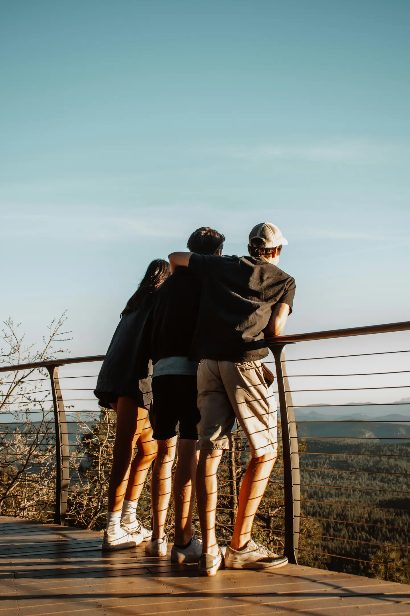 Three people stand on wooden deck leaning on railing, overlooking forested mountains under clear sky.