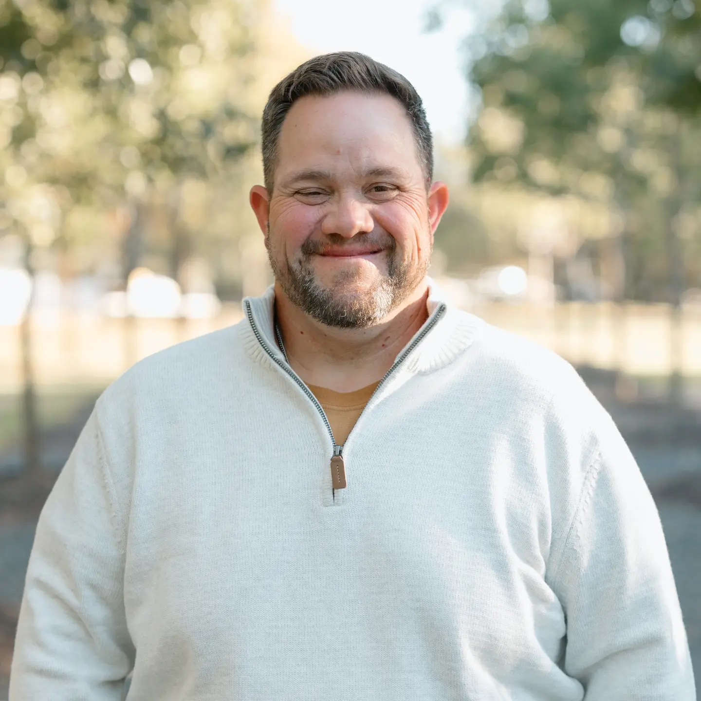 Smiling man wearing a white sweater outdoors with blurred trees in the background.