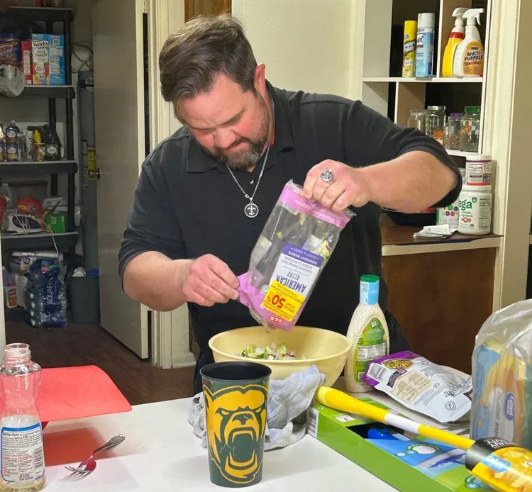 Man in black shirt pouring salad toppings from a plastic bag into a bowl in a kitchen.