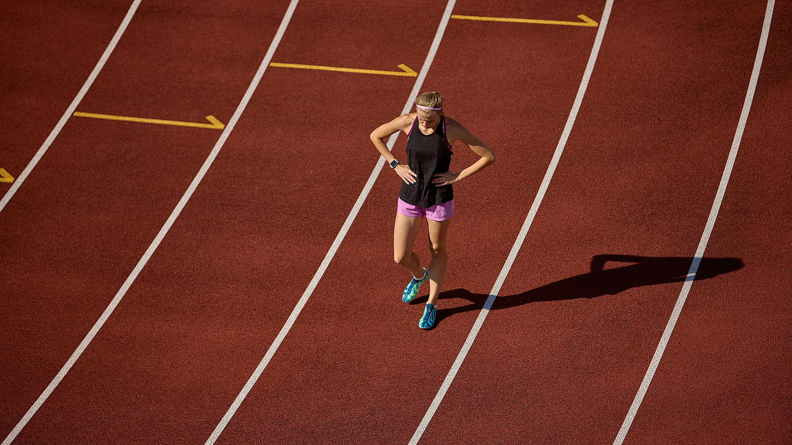 Athlete in black tank top and pink shorts resting on a red running track with lanes and arrows.