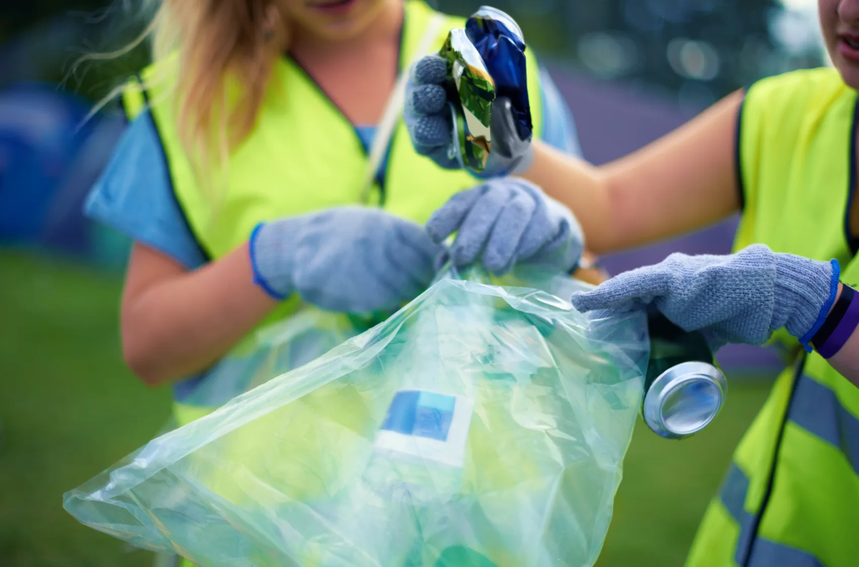 Volunteers in yellow vests wearing gloves sorting aluminum cans into a clear recycling bag outdoors.