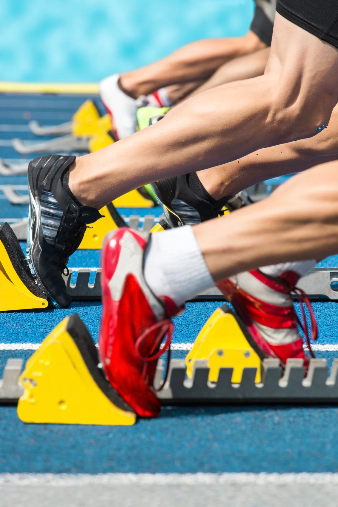 Closeup of athletes' legs and colorful running shoes on starting blocks on a blue track.
