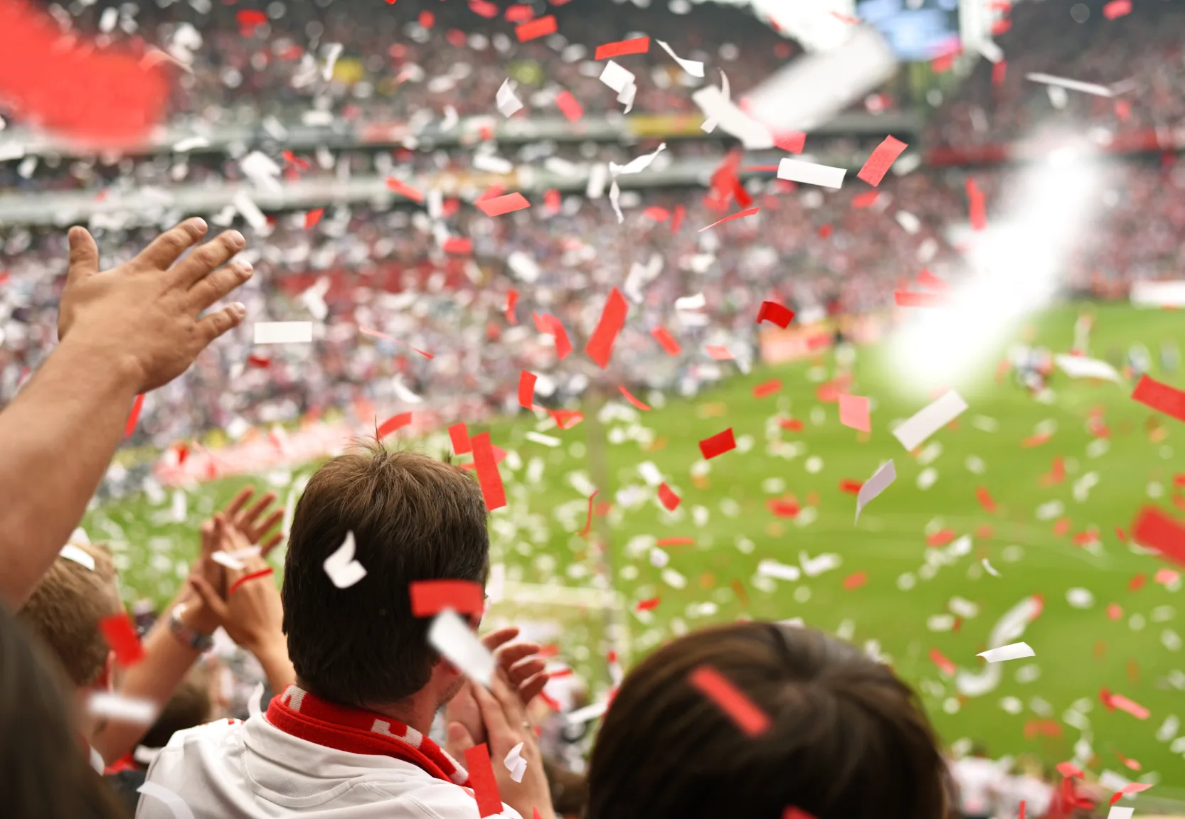 Fans in stadium cheering amid red and white confetti falling during a sports event.