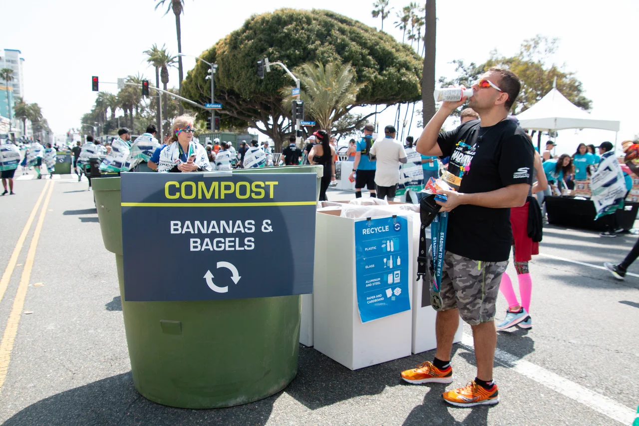 An LA Marathon runner drinking a beverage next to a recycle and compost station
