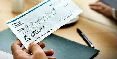 Hand holding a partially filled bank check with a pen on the desk in the background.