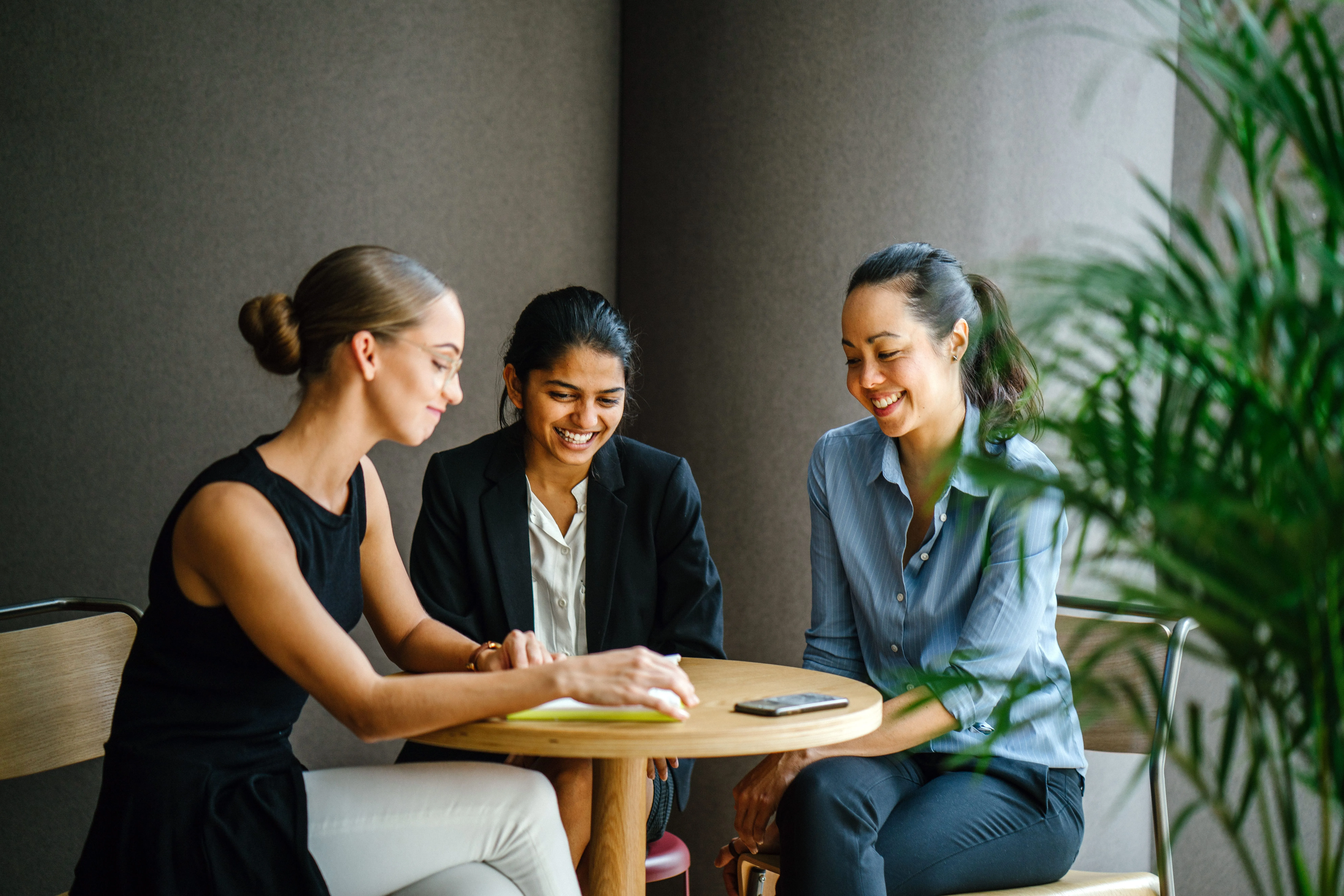 Three women sitting around a round wooden table, smiling and talking in a casual meeting.