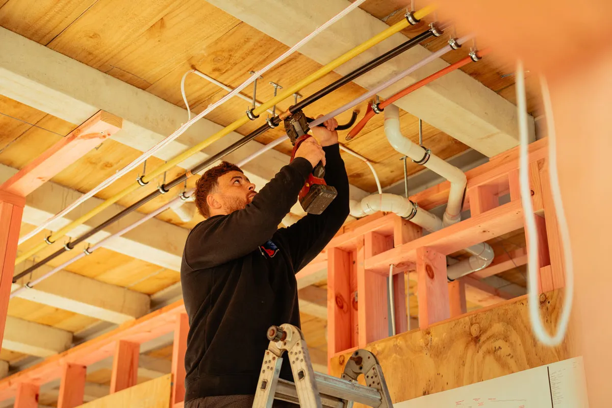 Man on ladder using a power tool to work on pipes attached to a wooden ceiling frame.