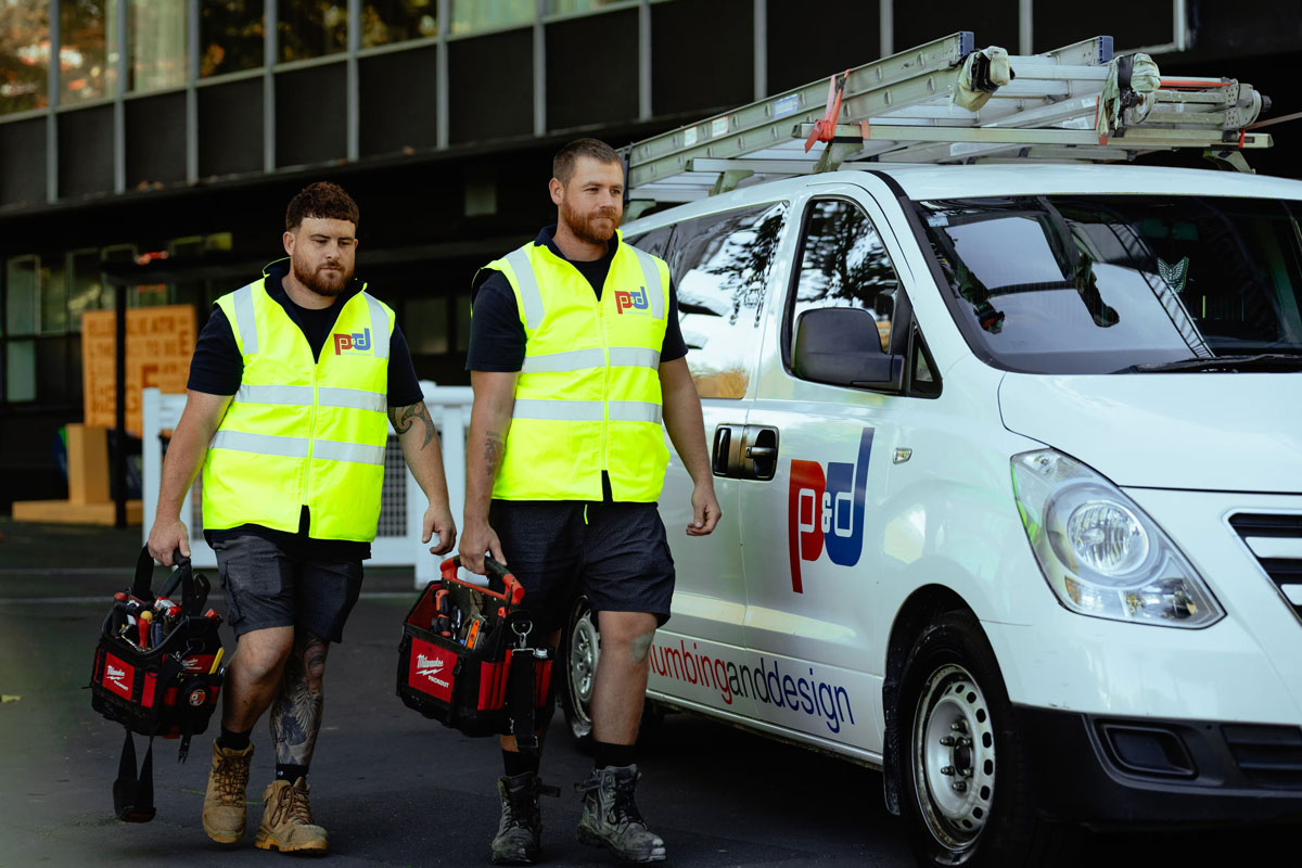 Two men in yellow safety vests walking with tool bags next to a white van with plumbing and design logo.