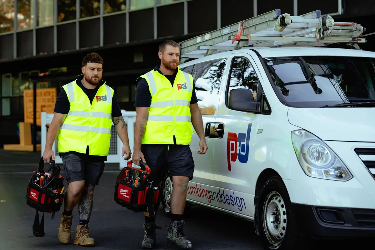 Two men in yellow safety vests walking with tool bags next to a white van with plumbing and design logo.