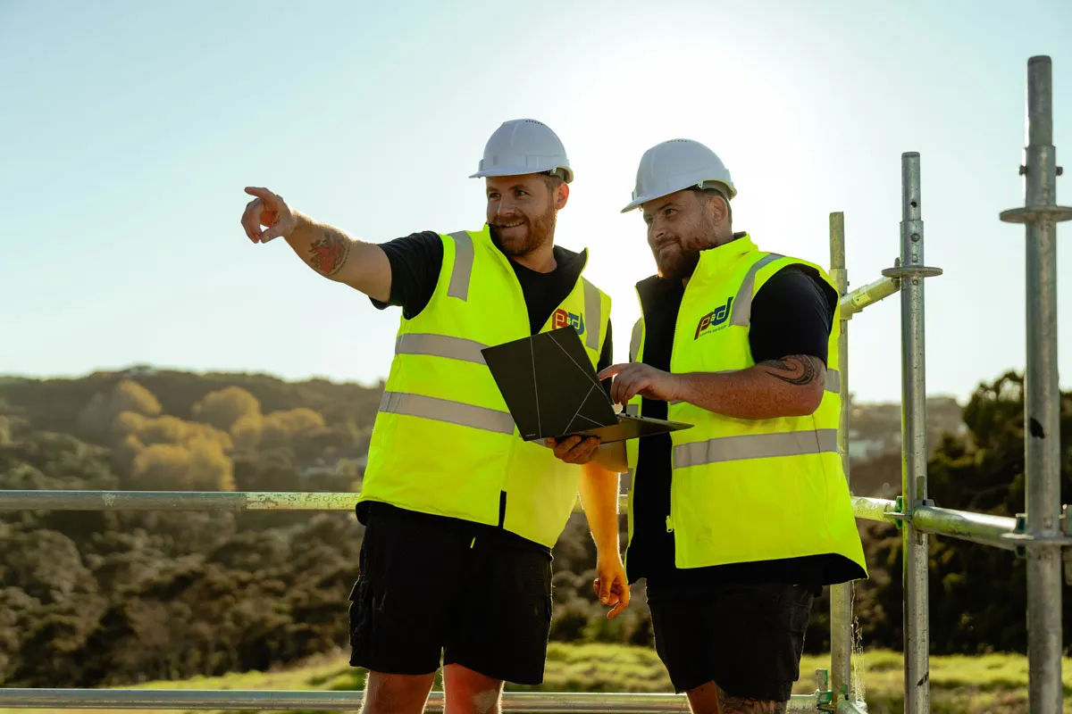 Two construction workers in helmets and yellow vests use a laptop outdoors near scaffolding.