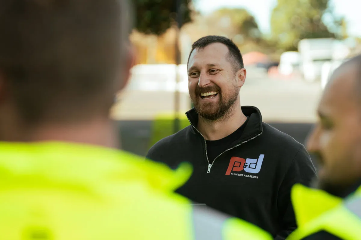 Smiling man in black P&D Plumbing and Design jacket talks with two men in yellow safety jackets.