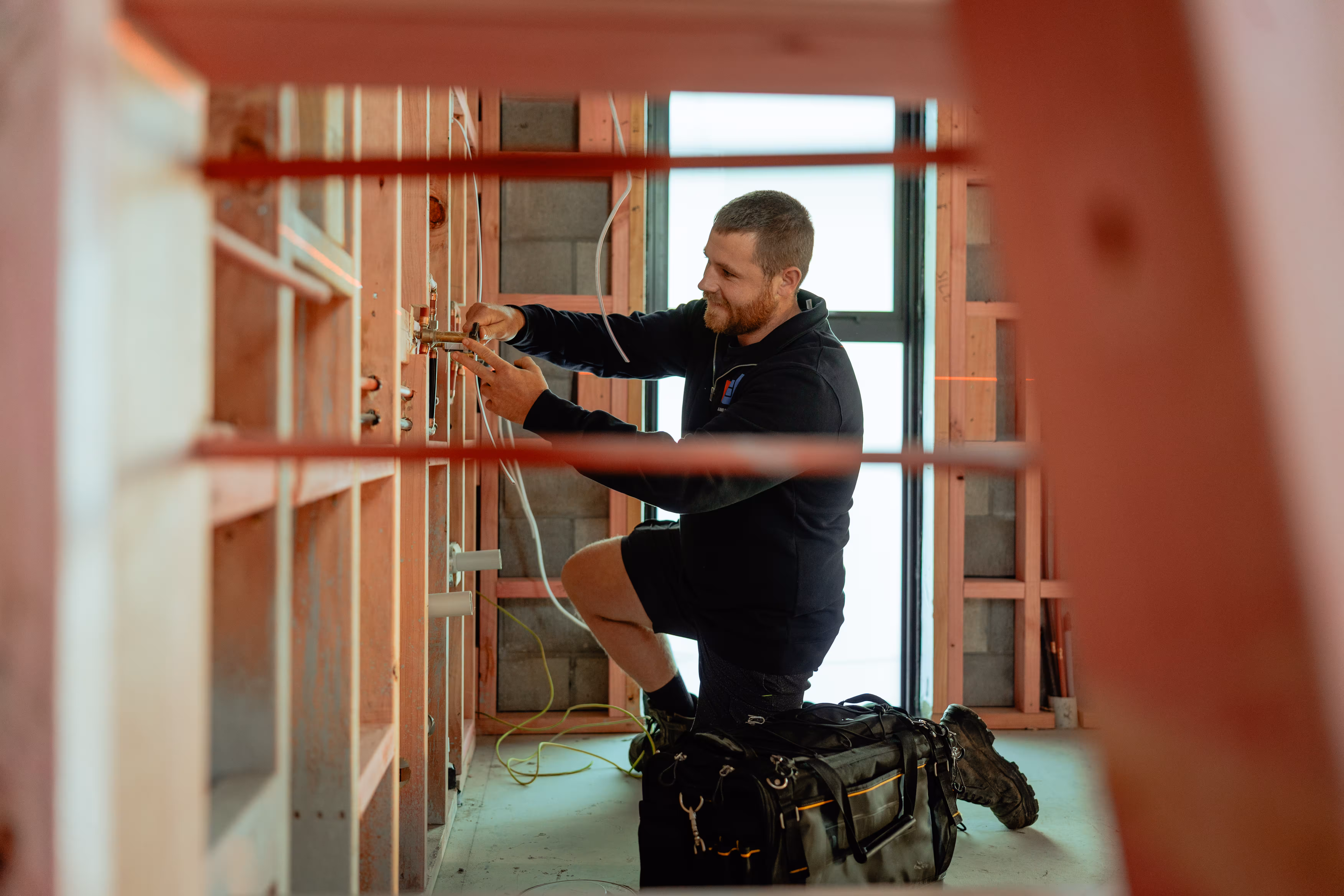 Man kneeling inside wooden frame house working with plumbing pipes and wires.