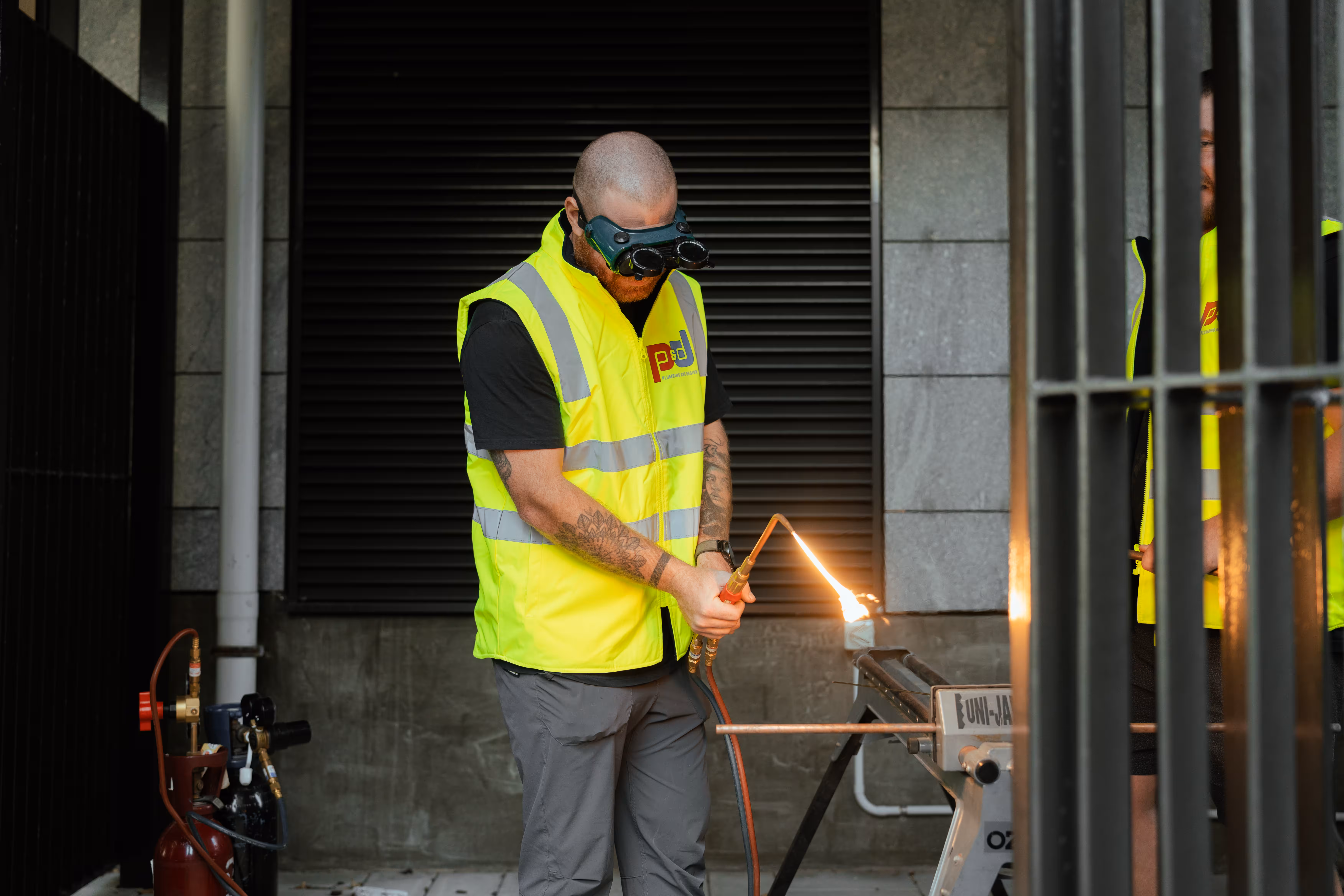 Worker in yellow safety vest using torch to weld or cut a pipe, wearing protective goggles.