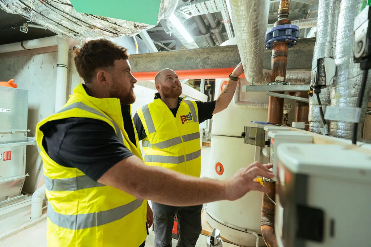 Two workers in yellow vests inspecting pipes and equipment in an industrial setting.