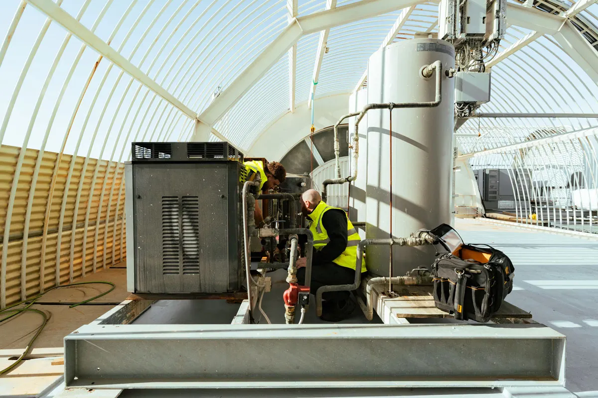 Two workers in yellow vests inspect and repair HVAC equipment inside a sunlit architectural structure.